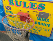 A carnival midway basketball hoop with a sign that says $2 and with rules. No cross throwing. Direct shots only. Must see shot.